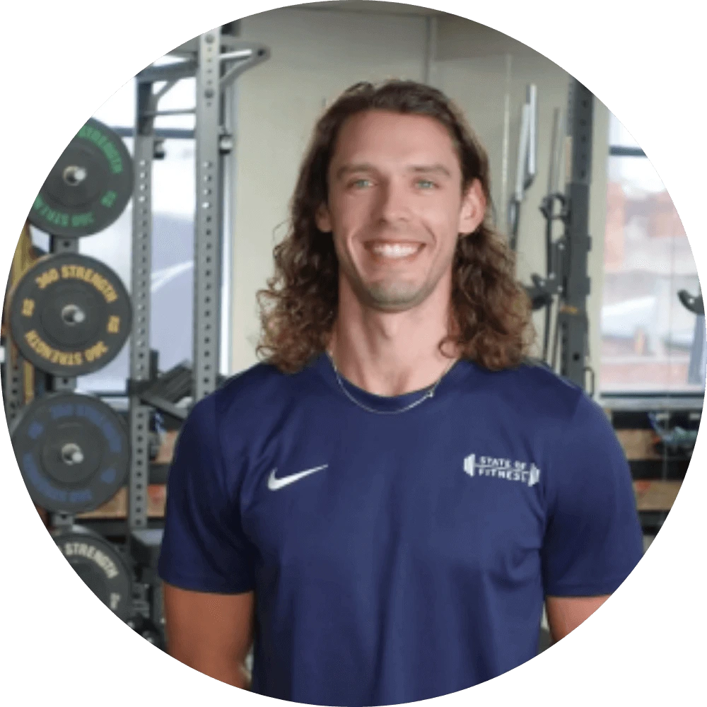 A man with long hair poses in front of gym equipment, ready to lead personal fitness sessions at the State of Fitness studio.