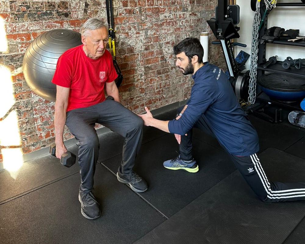 An elderly man performs a squat while a personal trainer provides support in a gym setting.