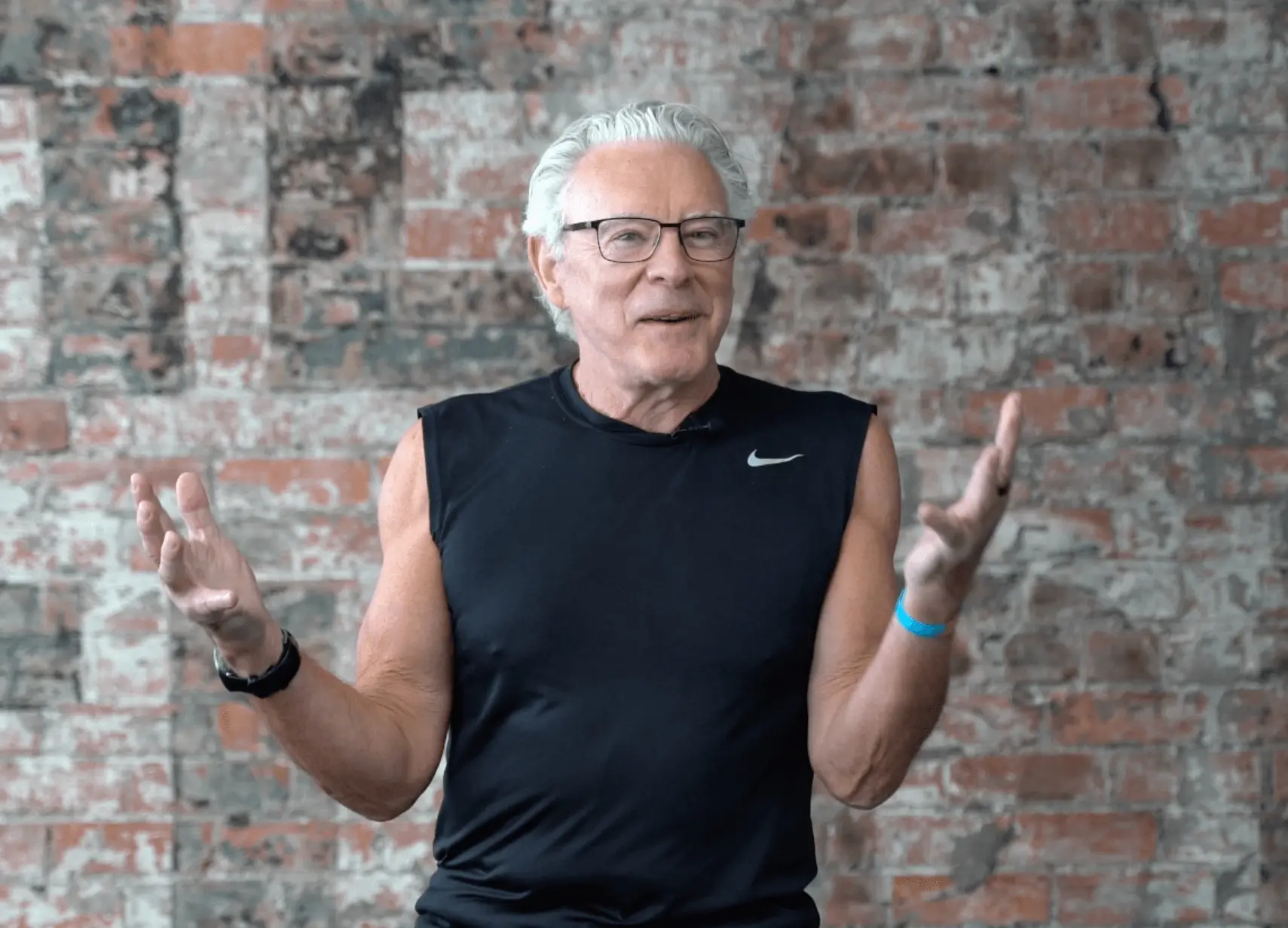 An enthusiastic older man with stylish glasses, dressed in dapper black gym wear, gestures expressively with his hands.