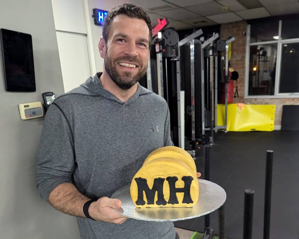 A man from the State of Fitness studio joyfully holds a cake decorated with the letters "MH" in celebration.