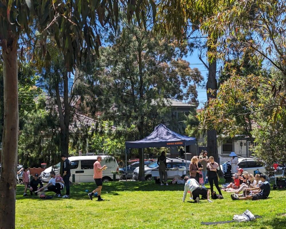 The State of Fitness studio team is sitting on the grass in a park, surrounded by trees and enjoying a sunny day together.